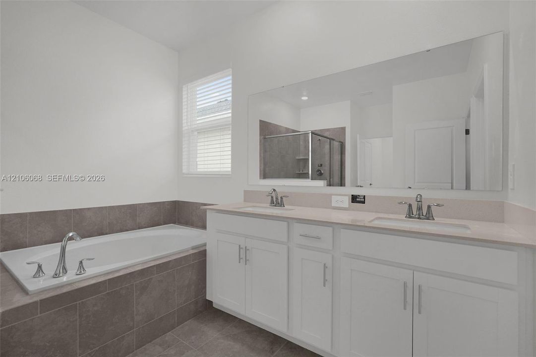 Dual-sink vanity with light-toned countertop, soaking tub featuring tile surround, oversized wall-mounted mirror, and tile flooring