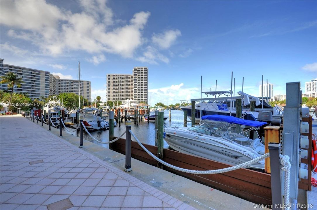 Waterfront boat slip featuring wood bulkhead, dockside pedestals, and rope railing with metal posts along a paved promenade