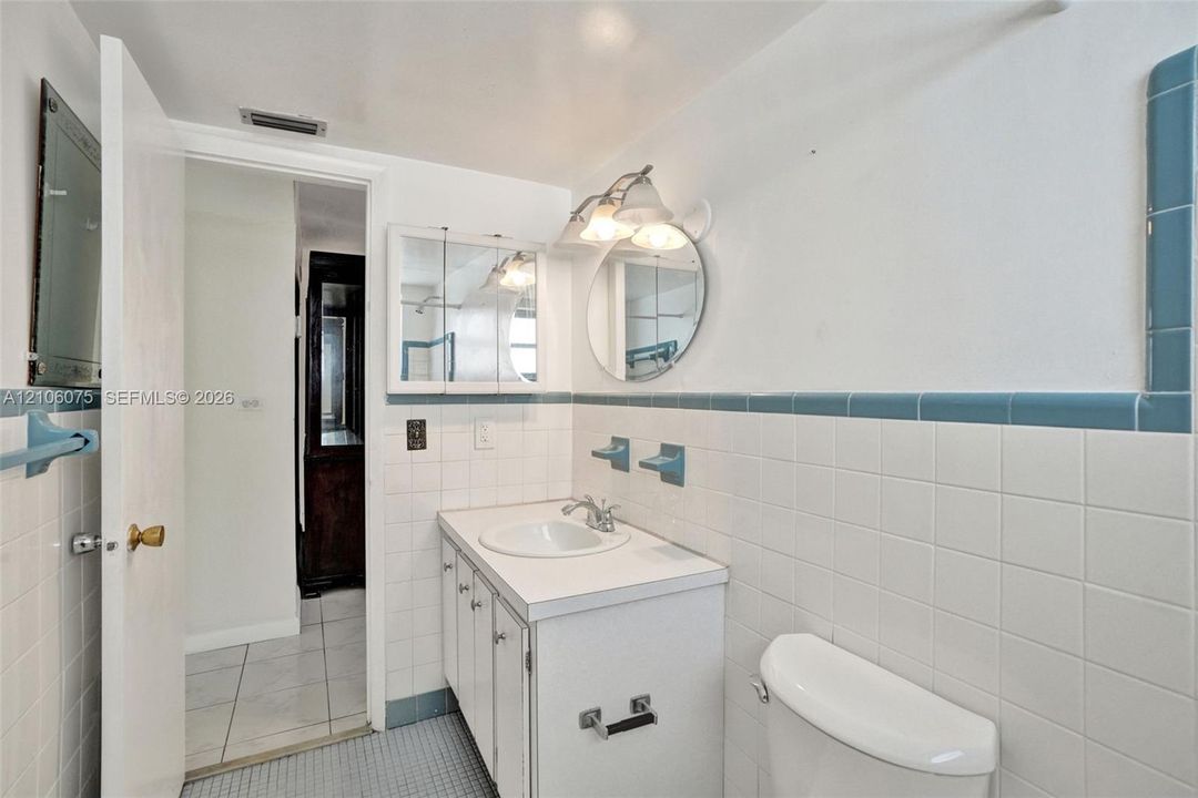 Bathroom featuring a white vanity cabinet with a integrated sink, ceramic wall tiling with blue trim, a round mirror, and a three-light fixture