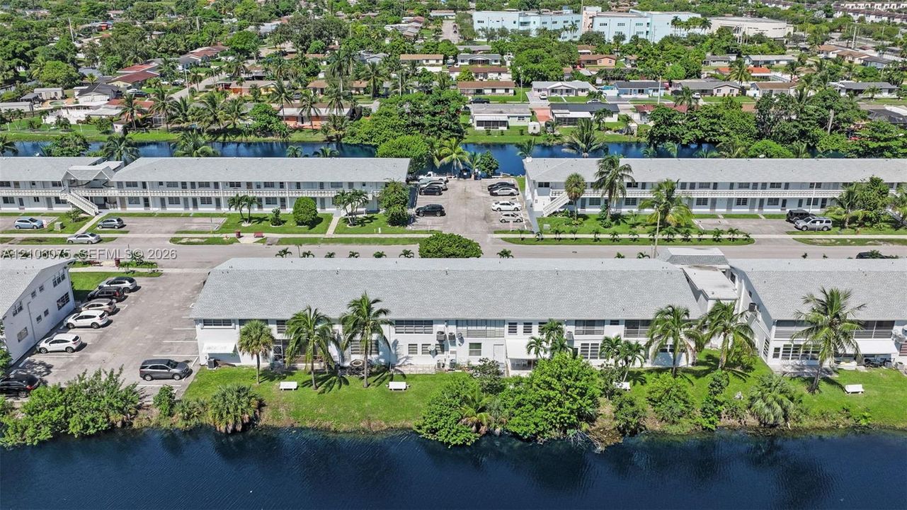 Aerial view of multi-unit residential complex featuring gray shingle roofing, paved surface parking, perimeter canal frontage, and mature palm trees