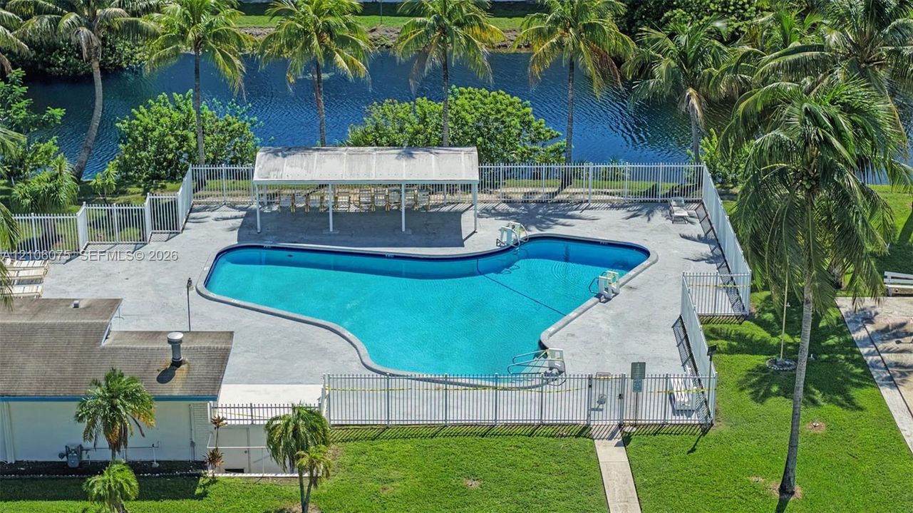 Outdoor swimming pool featuring a concrete deck, a covered patio structure, perimeter fencing, and mature palm trees along a canal