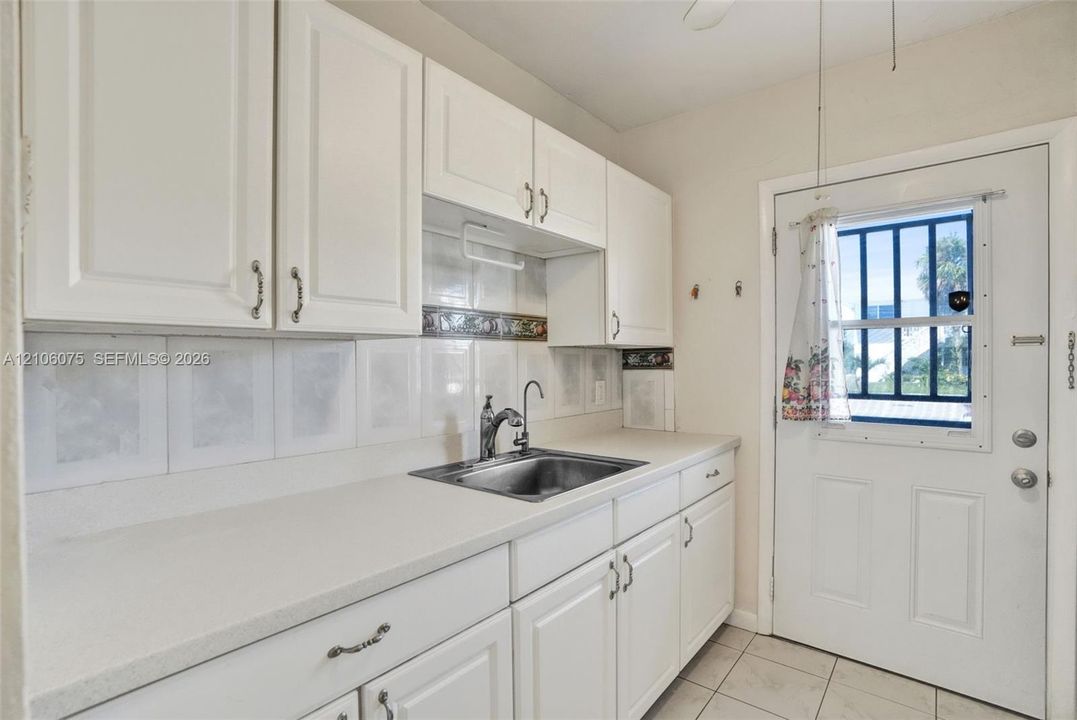 White cabinetry featuring integrated hardware, solid-surface countertops, tile backsplash, undermount stainless steel sink, and exterior door with inset window