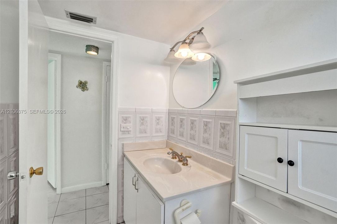 Vanity featuring a solid-surface countertop with an integrated basin, white built-in cabinetry, decorative tiled wall accents, a round wall-mounted mirror, and a three-light fixture