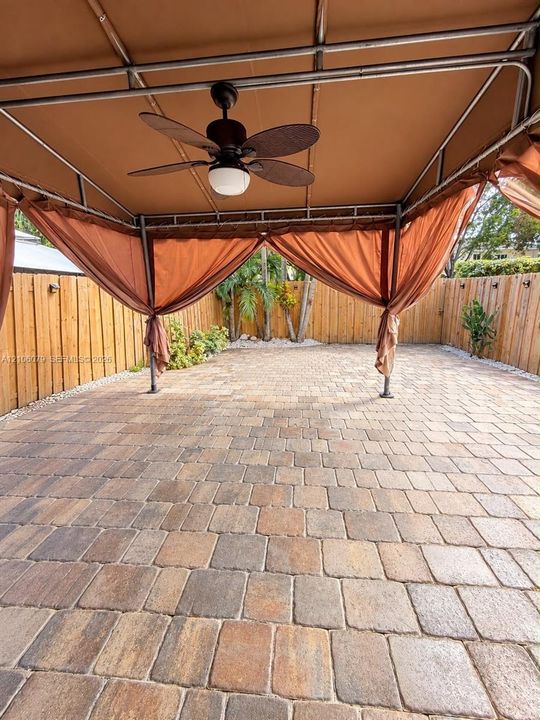 Outdoor patio featuring multi-tonal concrete pavers, a metal-framed overhead canopy, and a built-in ceiling fan, enclosed by a full-perimeter wood privacy fence