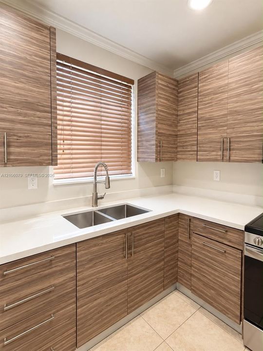 Kitchen featuring horizontal grain wood-finish cabinetry, white solid-surface countertops, dual-basin stainless steel sink with high-arc faucet, and tiled flooring