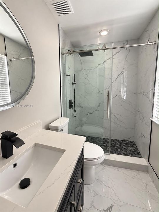 Dark-finish vanity with a light-toned countertop, integrated basin, matte black faucet, and frameless glass shower enclosure featuring large-format stone-patterned wall tile and a pebble-stone floor