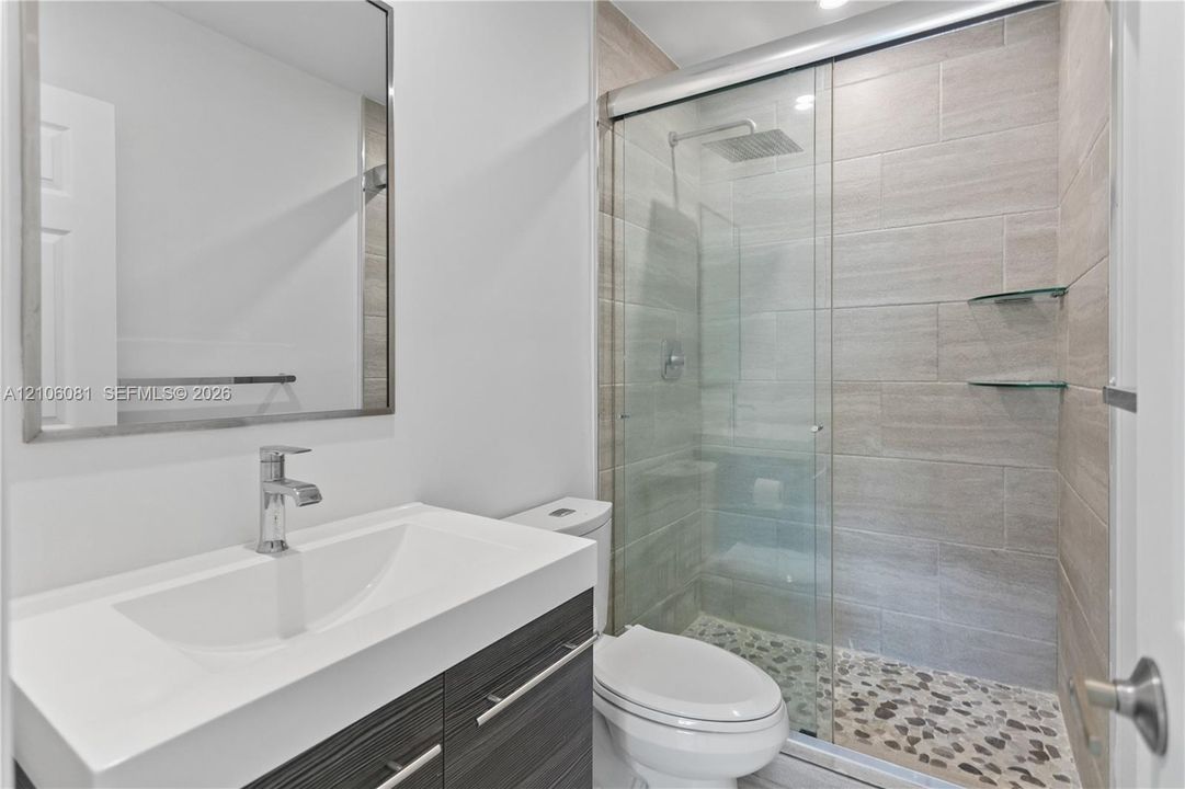Bathroom featuring a floating vanity with a white countertop, wall-mounted mirror, glass-enclosed shower with tile surround, pebble-stone flooring, and a rainfall showerhead