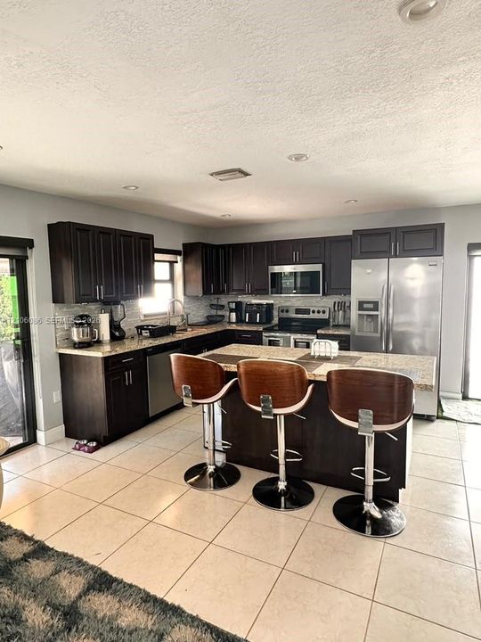 Kitchen featuring dark wood cabinetry, stone-finish countertops, stainless steel appliances, a central island, and ceramic tile flooring