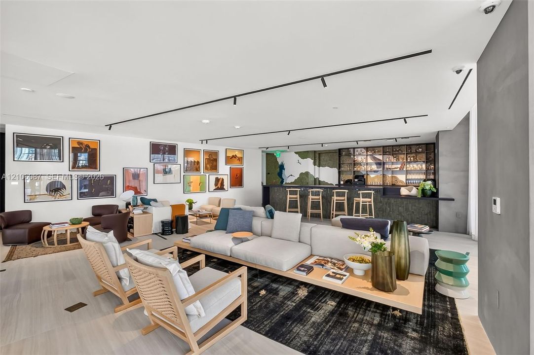 Open-concept space featuring light-toned wood-finish flooring, recessed track lighting, a built-in wet bar with dark stone-finish facing, and decorative glass shelving