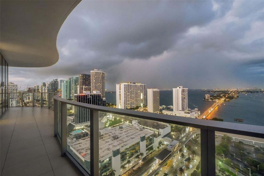 Curved balcony with large-format tile flooring and a glass-paneled railing overlooking an urban skyline and waterfront