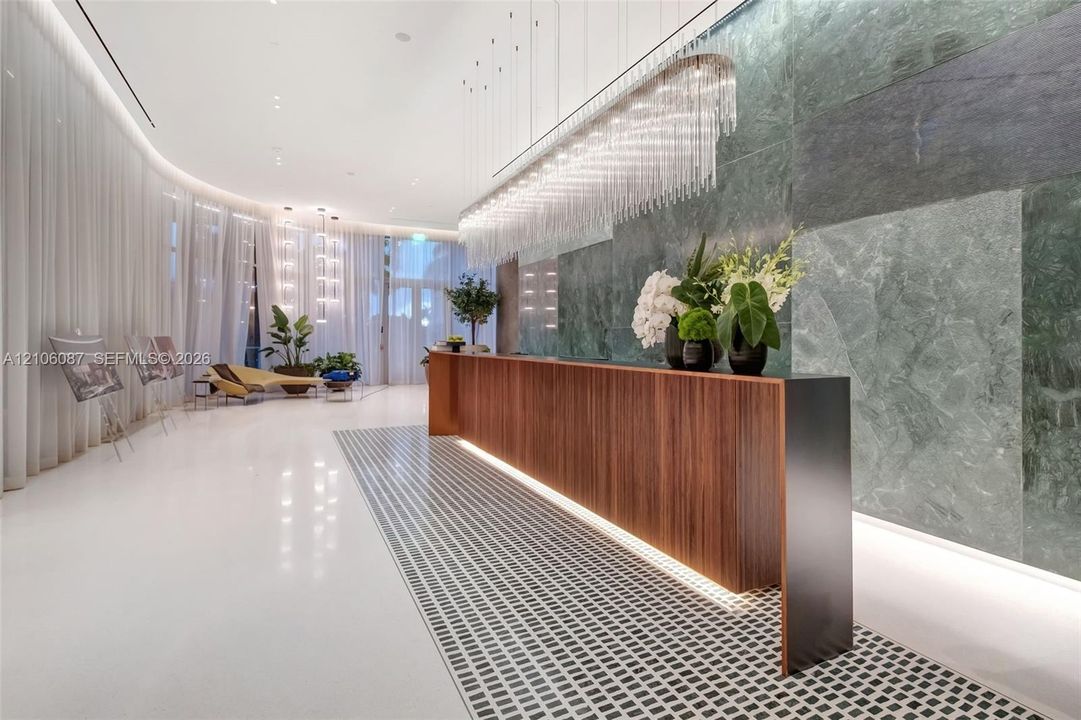 Reception area featuring a wood-panel console, large-format green stone wall cladding, custom crystal-style pendant lighting, polished white flooring, and inset mosaic floor detailing
