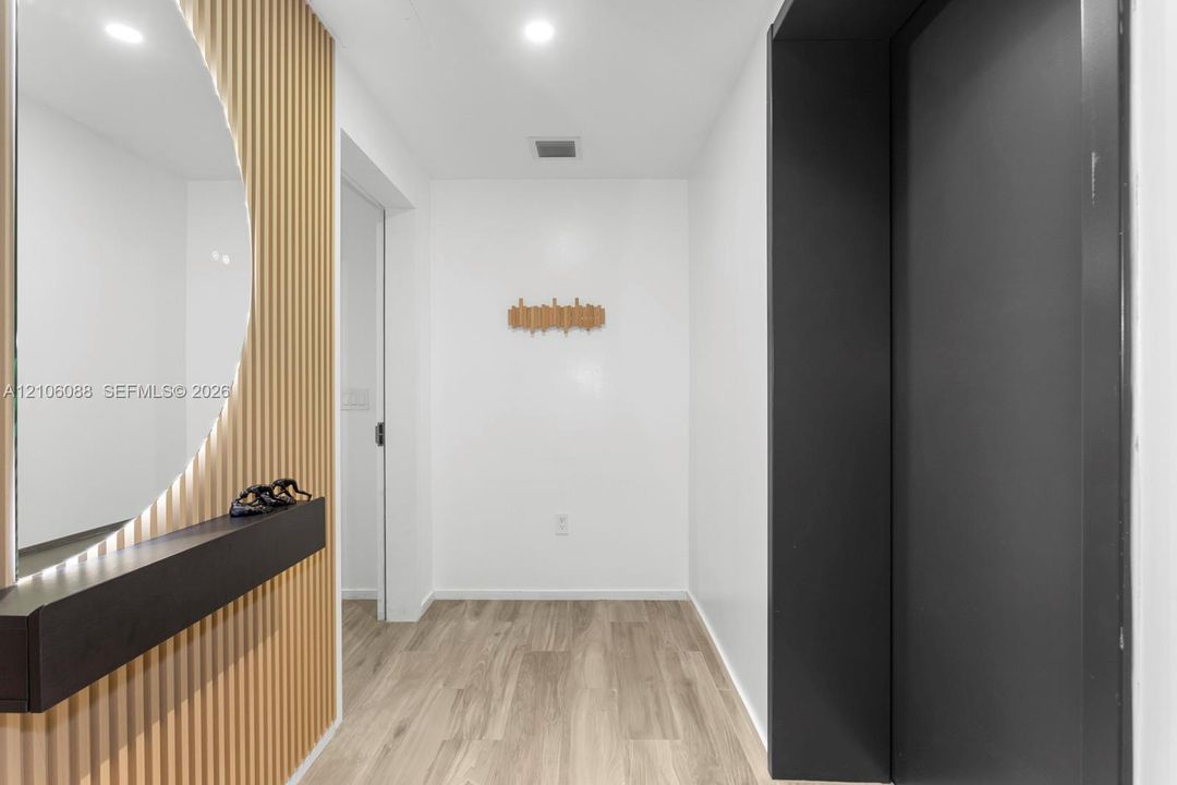 Foyer featuring wood-finish flooring, a vertical slat wood accent wall, a circular backlit wall mirror, and a sleek, matte black built-in floating shelf