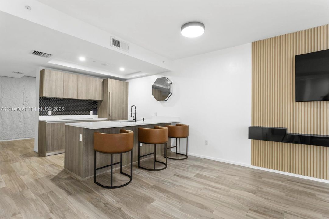 Kitchen featuring wood-finish cabinetry, white countertops, a dark hexagonal tile backsplash, a central island with an integrated faucet, and wood-finish flooring, complemented by a vertical slat wall accent