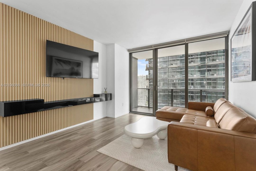 Living area featuring wood-finish flooring, a floor-to-ceiling slatted wood accent wall, a black wall-mounted floating console, and expansive floor-to-ceiling glass windows with sliding door access to a balcony