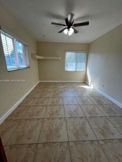 Neutral-toned interior space featuring ceramic tile flooring, a mounted ceiling fan with integrated lighting, two windows with horizontal blinds, and a built-in corner shelf