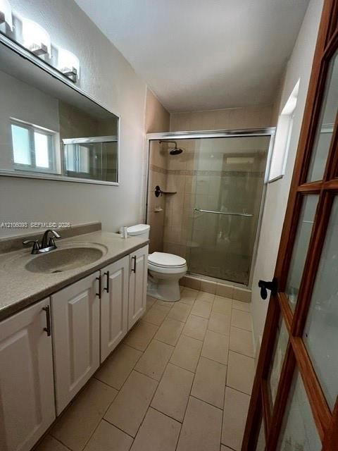 White cabinetry with dark hardware, stone-composite countertop, full-width vanity mirror, tile flooring, and walk-in shower with a sliding glass enclosure