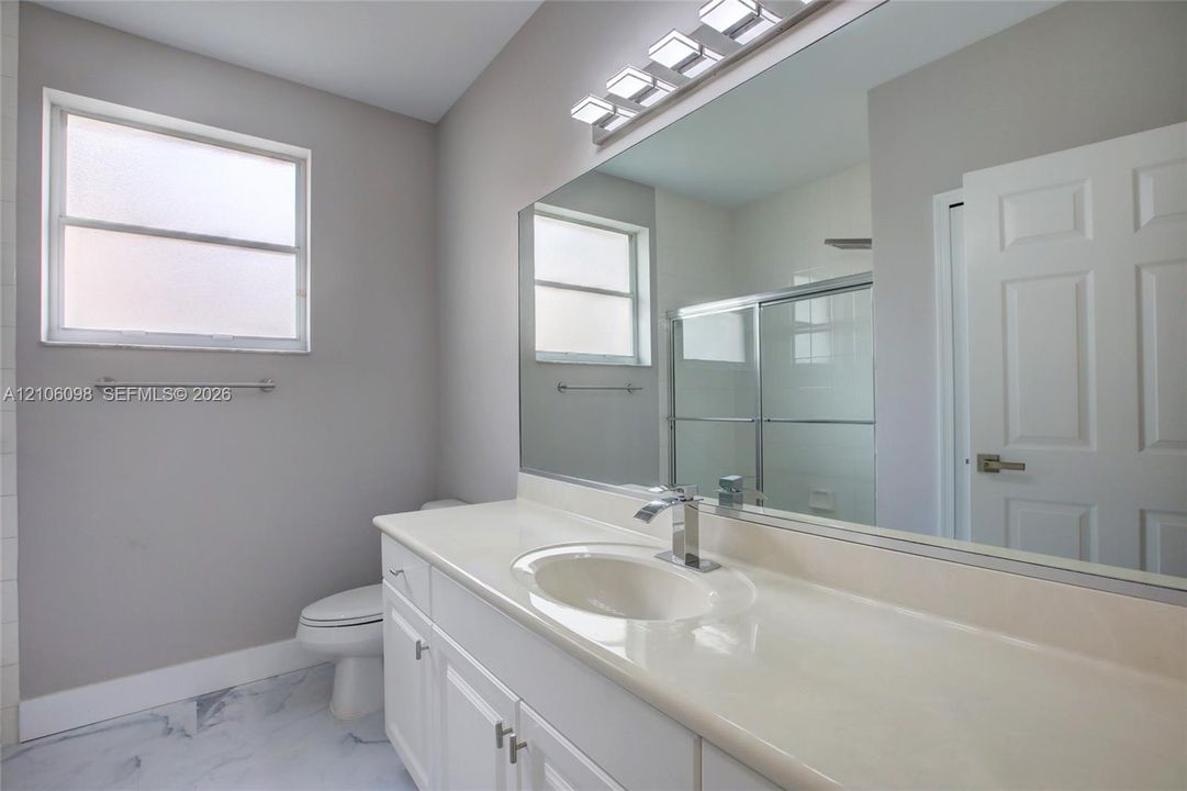 Bathroom featuring a solid-surface countertop with an integrated basin, white shaker-style vanity cabinetry, a full-width wall mirror, a multi-bulb light fixture, and a glass-enclosed shower