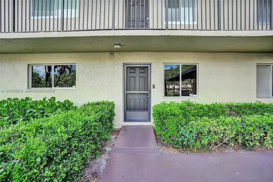 Stucco exterior featuring an entry door with a screen door, windows with aluminum frames, and a concrete walkway bordered by established shrubbery
