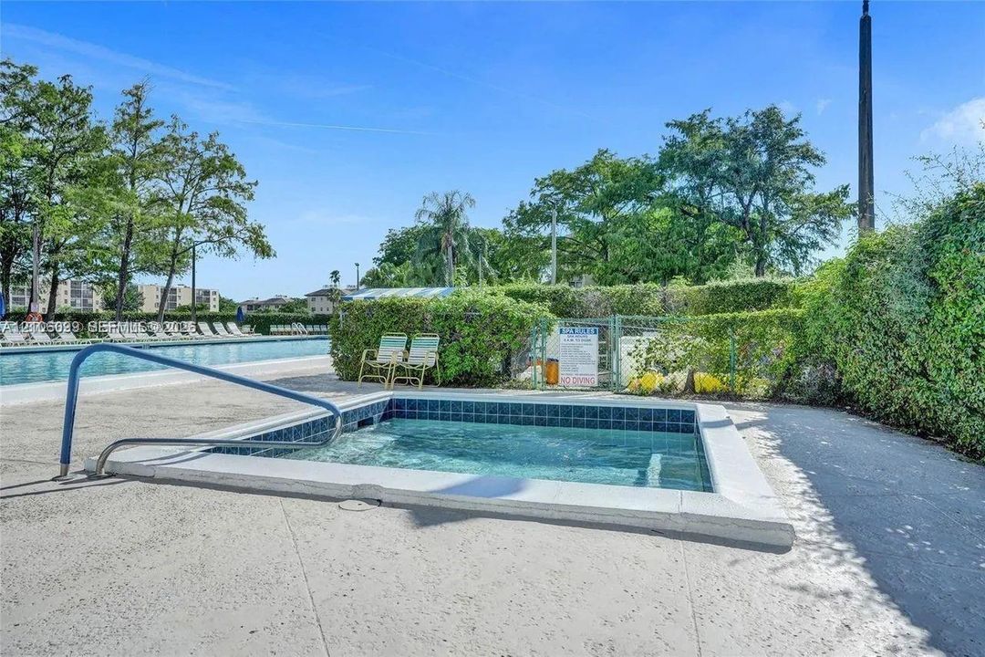 In-ground spa featuring blue tile trim and a metal handrail, situated on a concrete pool deck with manicured perimeter landscaping