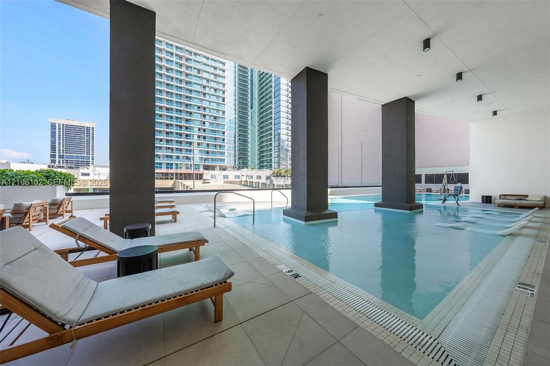 Covered swimming pool featuring large structural columns, light-toned tile decking, and recessed ceiling lighting