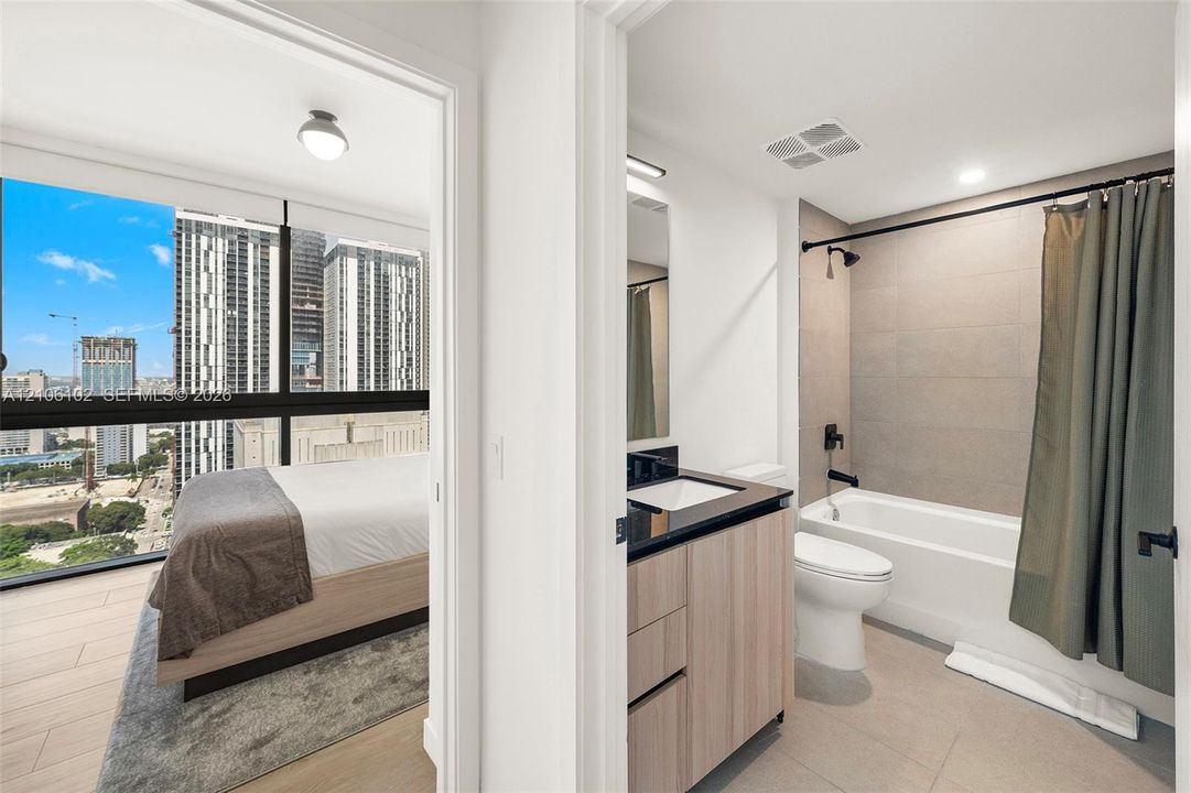 Bathroom featuring a wood-finish vanity, dark stone countertop, undermount sink, and full-height wall tiling in the shower area, paired with an room featuring floor-to-ceiling windows and wood-finish flooring