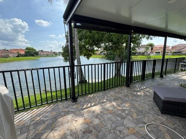 Covered patio featuring a flagstone-patterned paved floor and black metal railings, overlooking a lake with mature landscaping