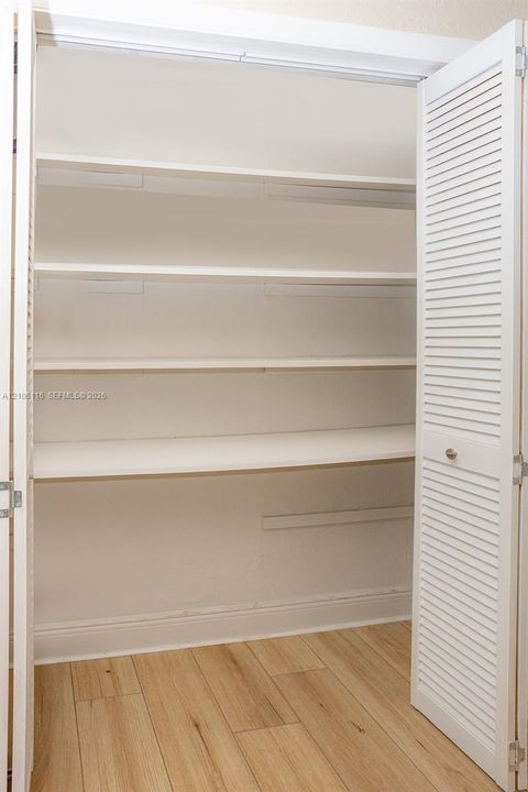 Built-in storage closet featuring white louvered doors and four interior shelves over wood-finish flooring