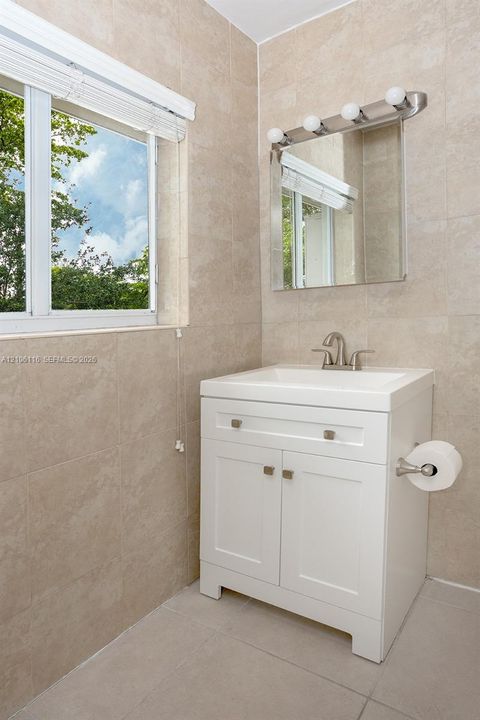 White vanity with dual-door cabinetry and integrated sink, polished metal faucet, wall-mounted light fixture above a square mirror, and full-height tile wall surround