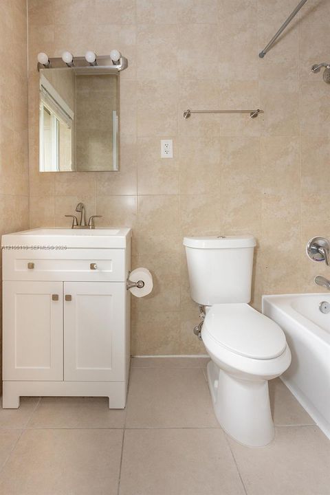 Bathroom featuring floor-to-ceiling ceramic wall tiling, white shaker-style vanity cabinet with a squared basin, brushed-nickel vanity light fixture, and a standard bathtub with an integrated shower head
