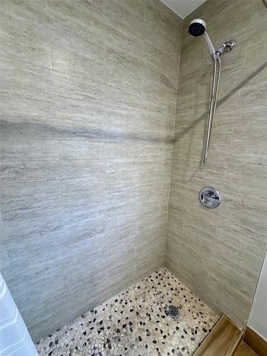 Shower featuring horizontal stone-textured wall tile, pebble-mosaic flooring, and a chrome hand-held showerhead with integrated wall-mounted pressure control