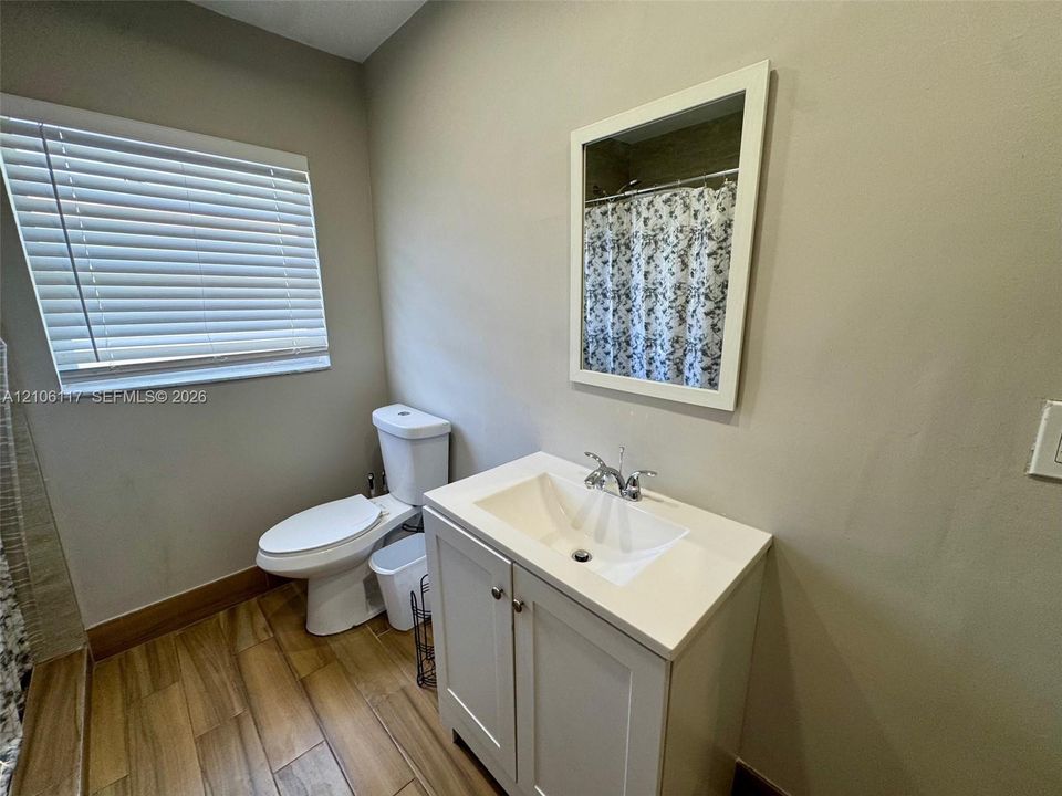 Bathroom featuring a white vanity cabinet with a stone-top basin, wood-finish flooring, a wall-mounted framed mirror, and a window with horizontal blinds
