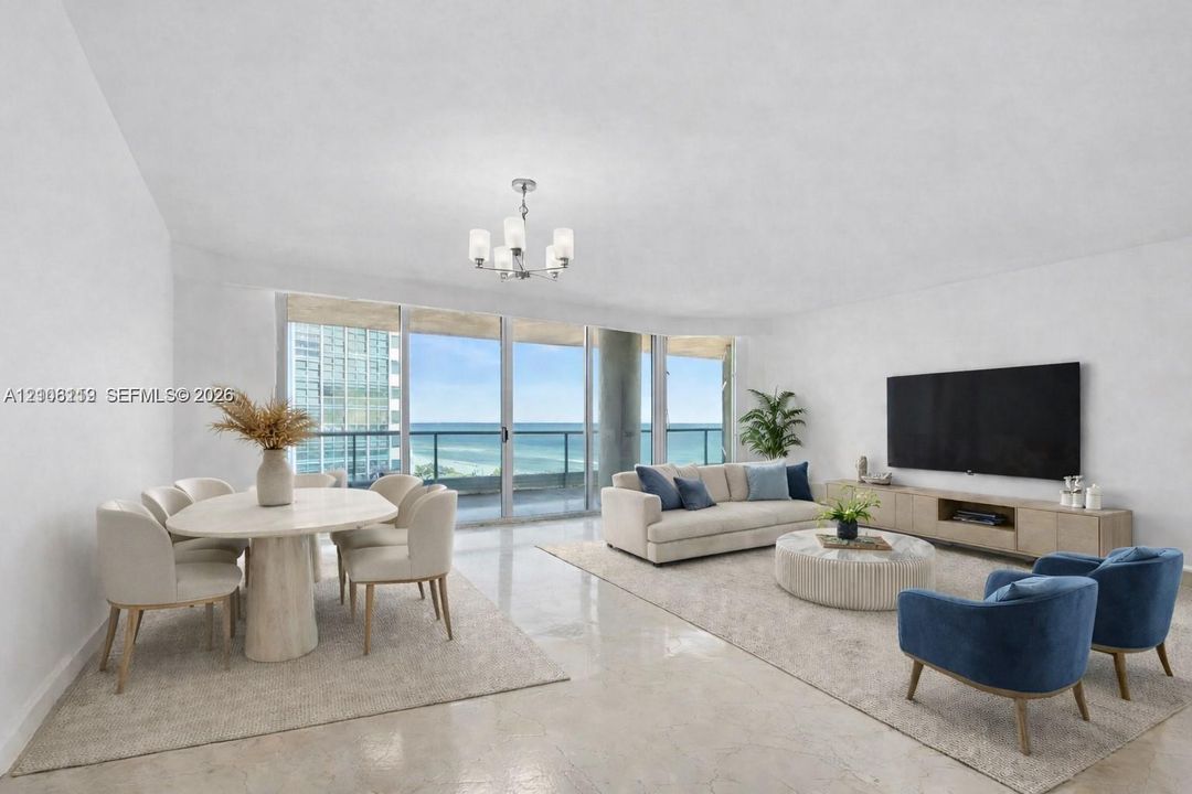 Open-concept interior featuring light-toned stone flooring, floor-to-ceiling glass sliding doors, a glass-railed balcony, and a multi-arm chandelier