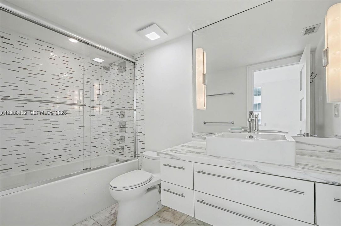 Bathroom featuring a white vanity with a stone-finish countertop and square vessel sink, full-width wall mirror with integrated lighting, glass-enclosed shower and tub combination, and decorative mosaic tile shower wall