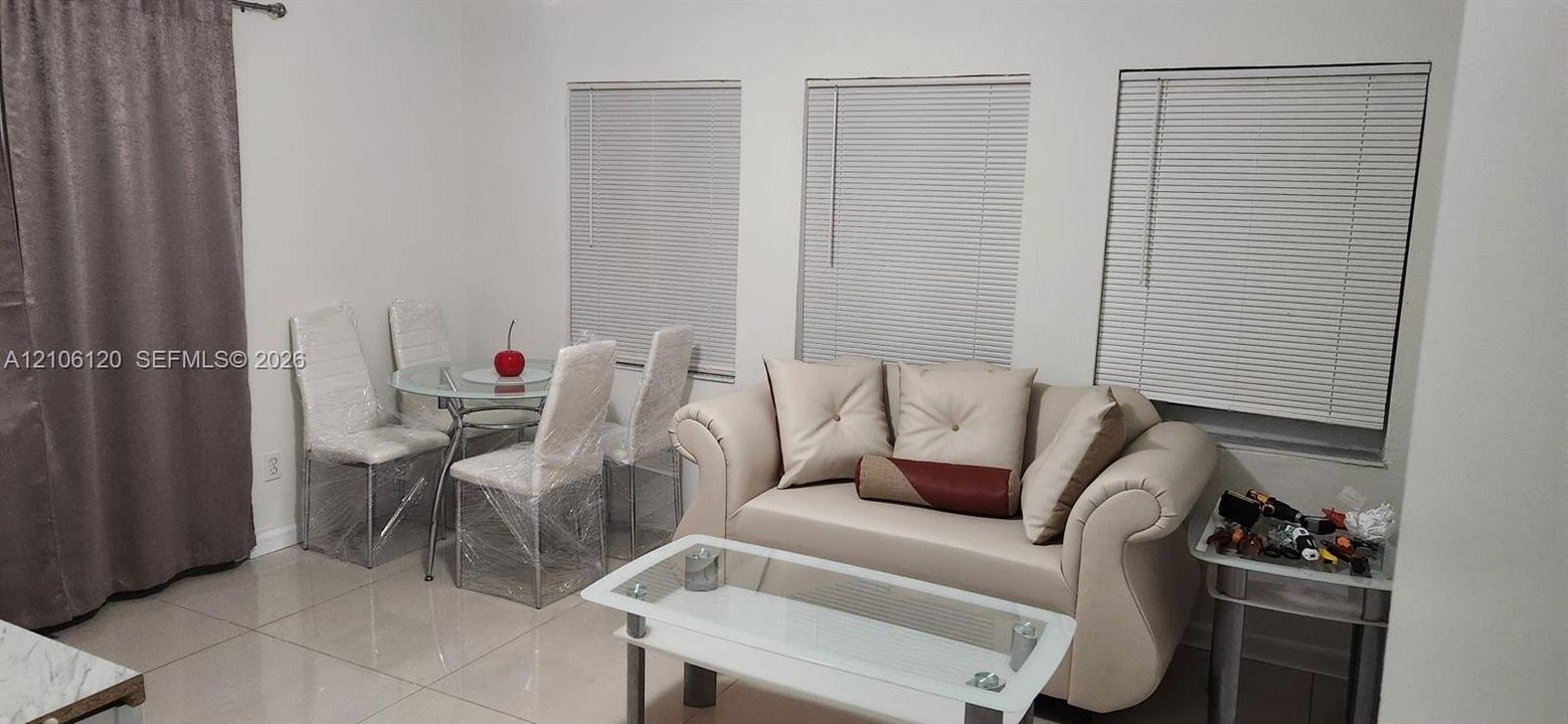 Neutral-toned tile flooring, three large windows featuring horizontal blinds, and floor-to-ceiling drapery