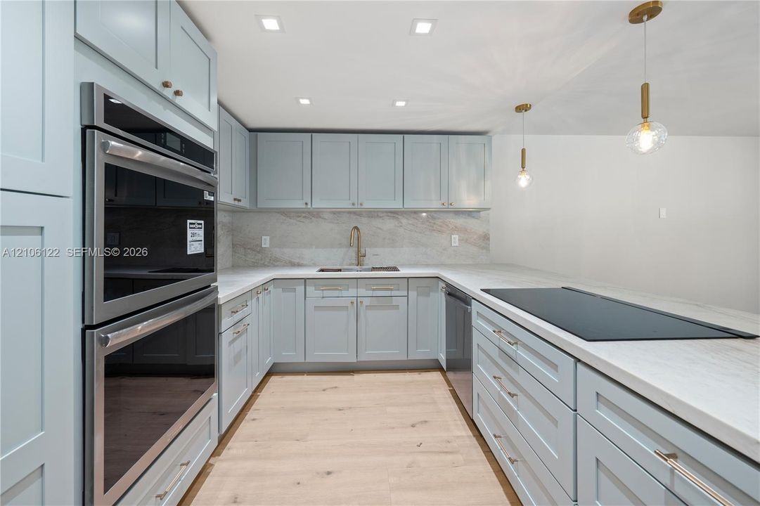 Kitchen featuring light-toned shaker cabinetry, stone-finish countertops and backsplash, integrated double wall ovens, wood-finish flooring, and pendant lighting