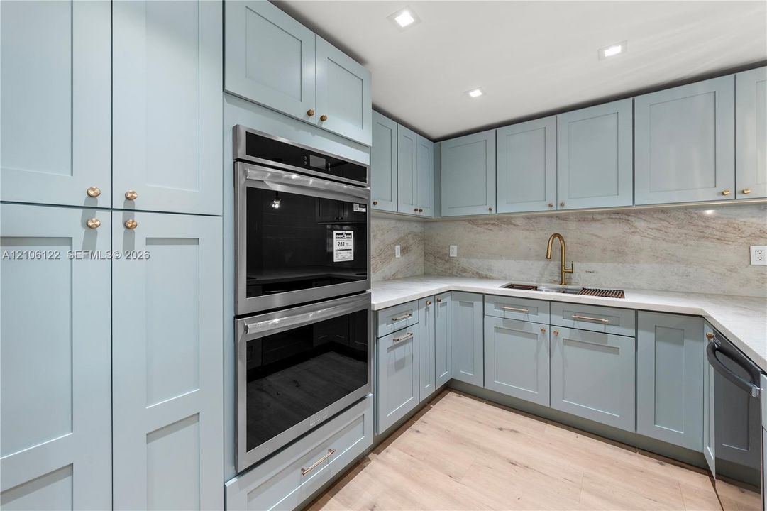 Kitchen featuring custom soft-blue shaker cabinetry, stone backsplash, built-in double wall oven, gold-finish faucet, and wood-finish flooring