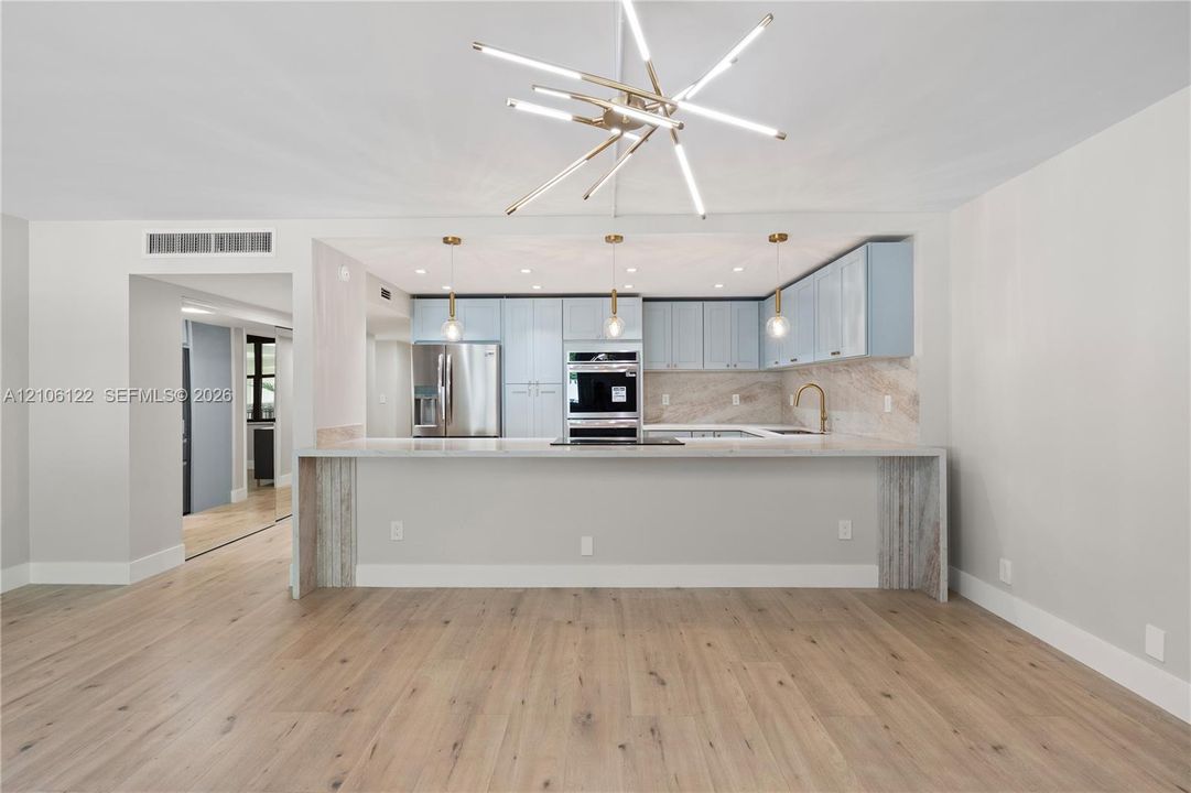 Kitchen featuring light blue cabinetry, stone-finish backsplash, wood-finish flooring, a waterfall-edge peninsula with stone countertop, and contemporary linear geometric chandelier