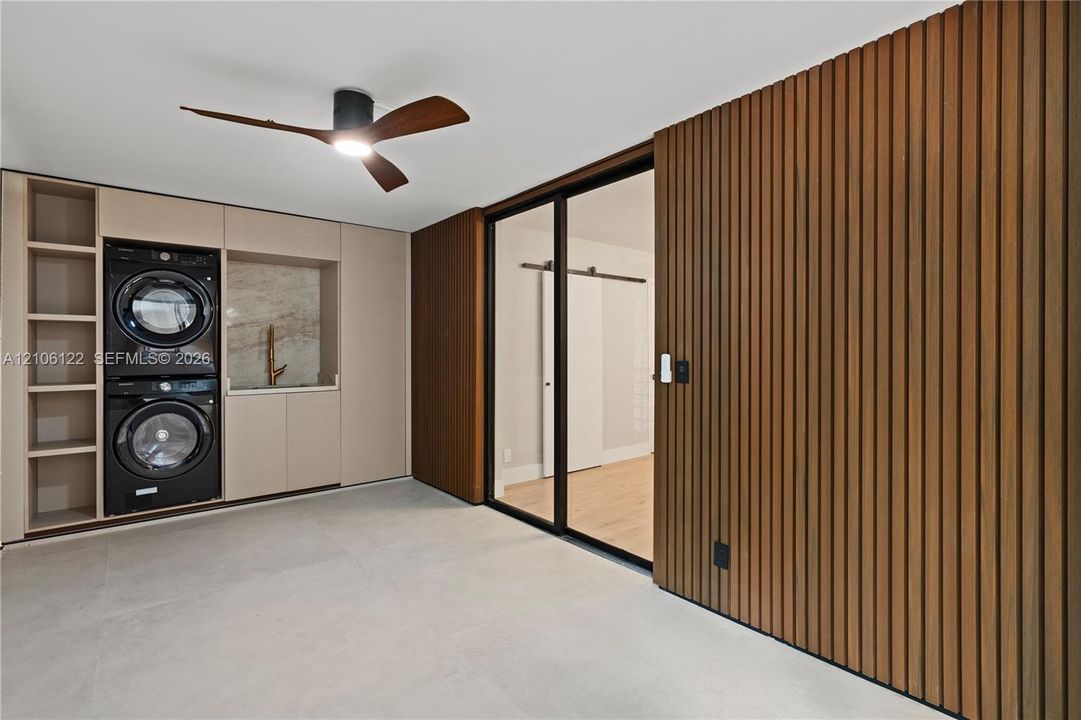 Built-in cabinetry with integrated appliance housing, vertical wood-slat wall paneling, full-height glass-paneled door with black frame, matte-finish flooring, and minimalist ceiling fan with integrated lighting