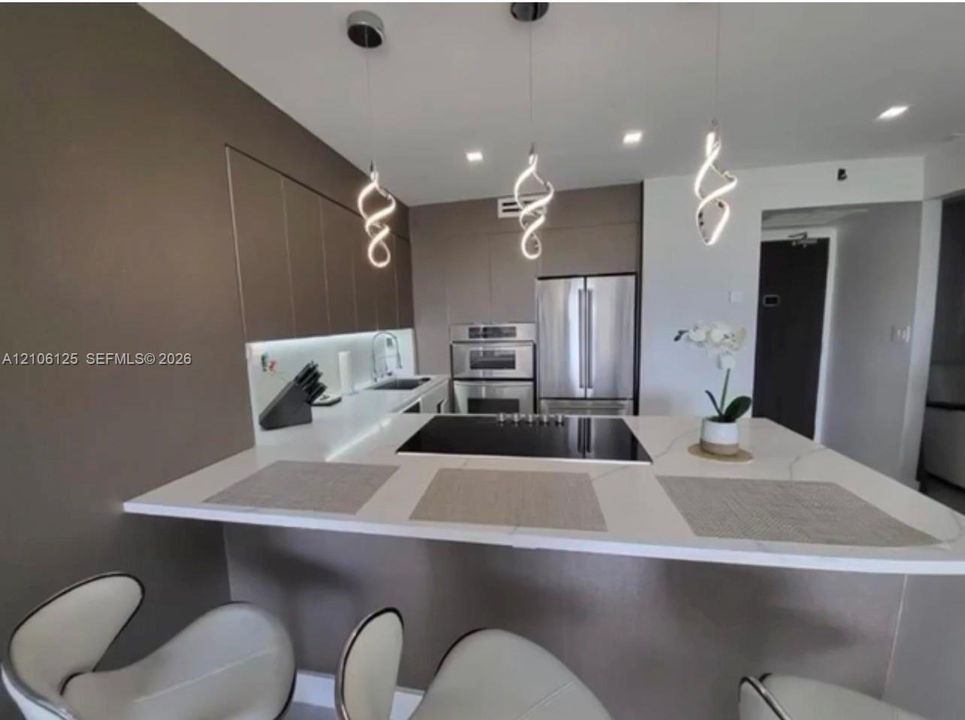 Contemporary kitchen featuring a spacious quartz peninsula, stainless steel double wall ovens, a flat-surface cooktop, matte-finish cabinetry, and sculptural pendant lighting