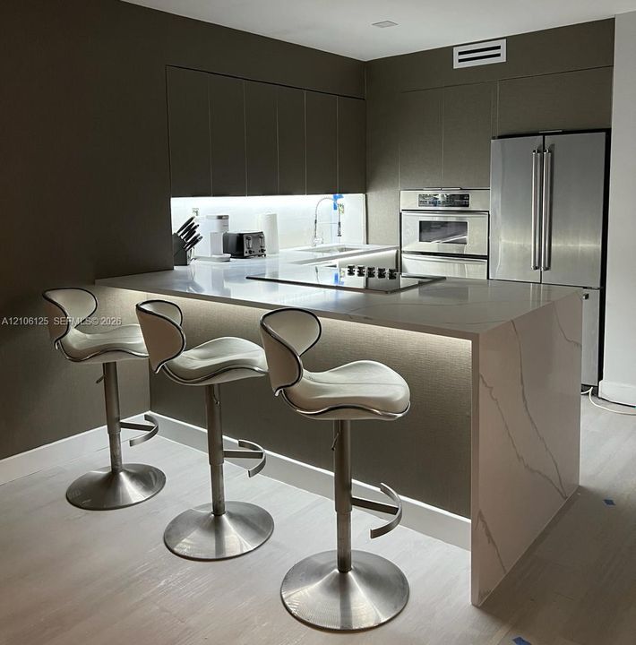 Contemporary kitchen featuring flat-panel cabinetry, a waterfall-edge countertop, an integrated cooktop, and a stainless steel wall oven and refrigerator
