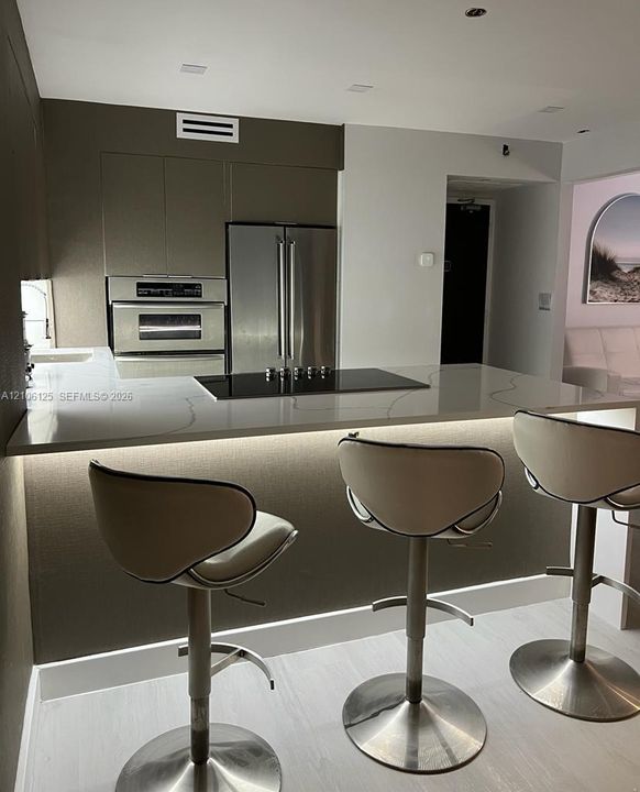 Kitchen featuring flat-panel cabinetry, stainless steel appliances including a wall-mounted oven, and a solid-surface waterfall island with an integrated cooktop and under-counter lighting