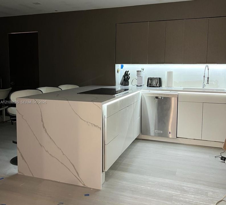 Contemporary kitchen featuring a stone waterfall island with an integrated cooktop, flat-panel cabinetry, stainless steel dishwasher, and under-cabinet accent lighting