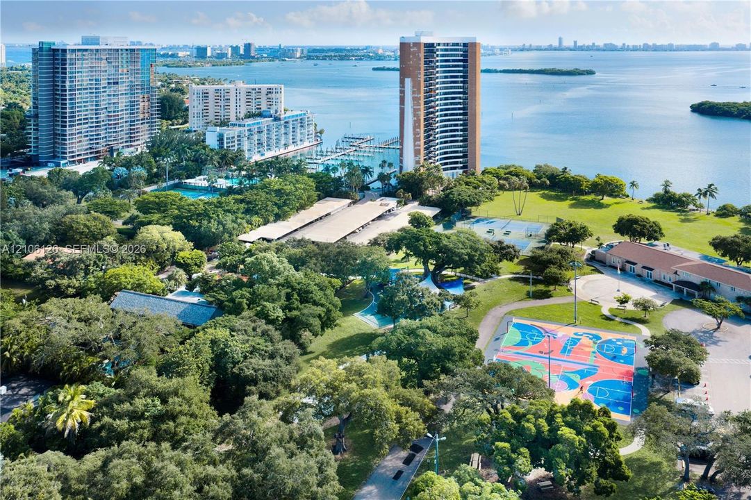 Aerial perspective showcasing waterfront grounds featuring mature tree canopy, multi-colored outdoor sports court, open lawn space, and proximity to marina infrastructure