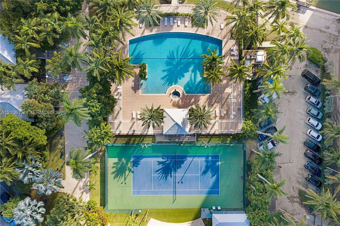 Aerial view of recreational amenities featuring a paved swimming pool deck, integrated spa, hard-surface tennis court, and mature palm landscaping