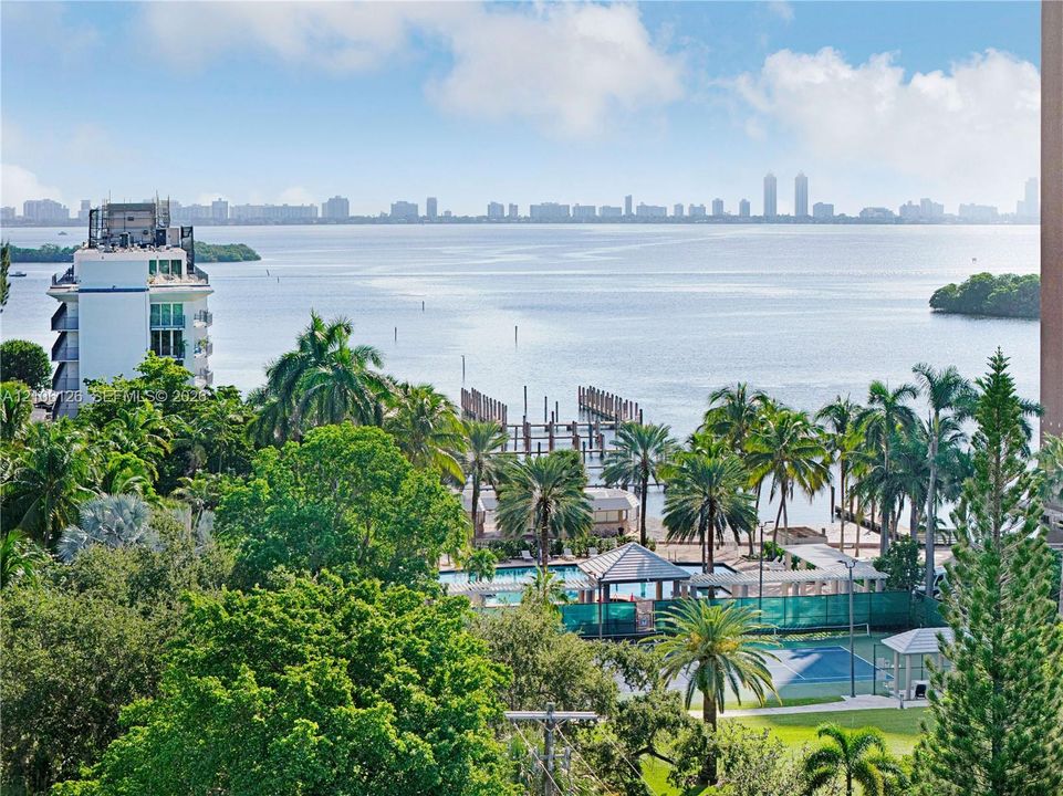 Panoramic water and skyline backdrop featuring mature tropical landscaping, an outdoor swimming pool, and a tennis court