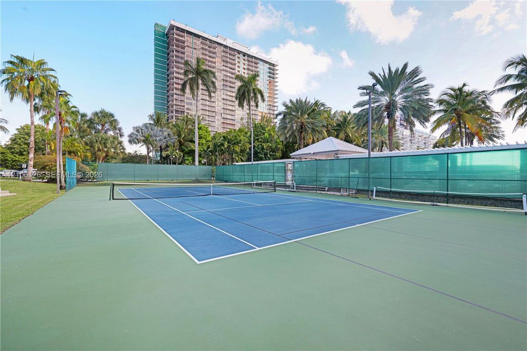 Hard-surfaced tennis court featuring a blue and green color scheme, perimeter fencing, and surrounding mature palm trees