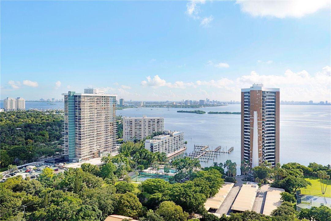 Aerial perspective showcasing a coastal cityscape featuring multi-story high-rise structures, waterfront access, a boat dock, and surrounding mature vegetation