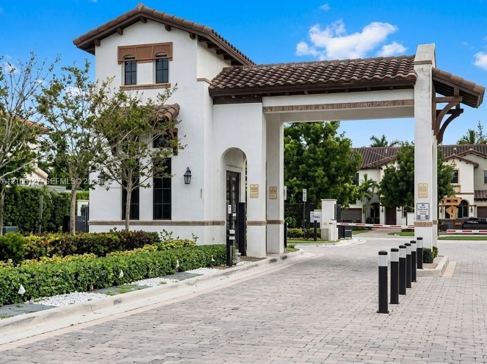 Gated entrance featuring a stucco guardhouse, clay tile roof, paver driveway, and controlled-access barrier arm