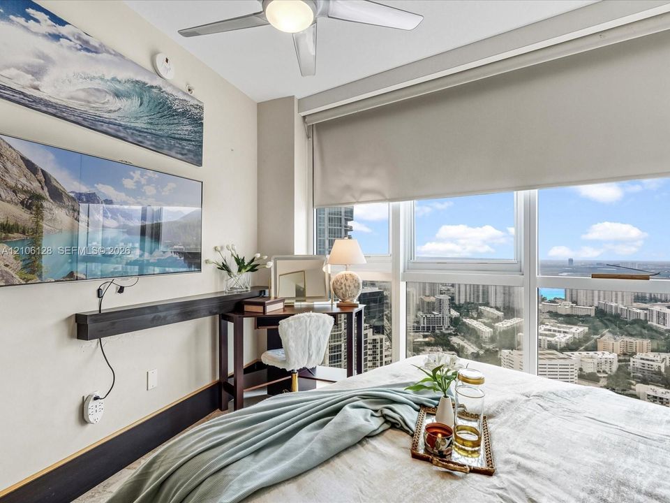 Floor-to-ceiling windows with urban skyline backdrop, custom wall-mounted shelving, overhead ceiling fan, and roller shades