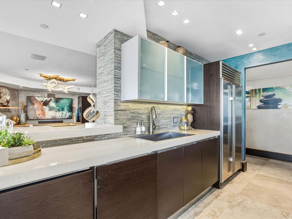 Modern kitchen featuring dark wood-finish cabinetry, frosted glass upper cabinets, a light-toned countertop, a built-in stainless steel refrigerator, and a textured stacked-stone backsplash